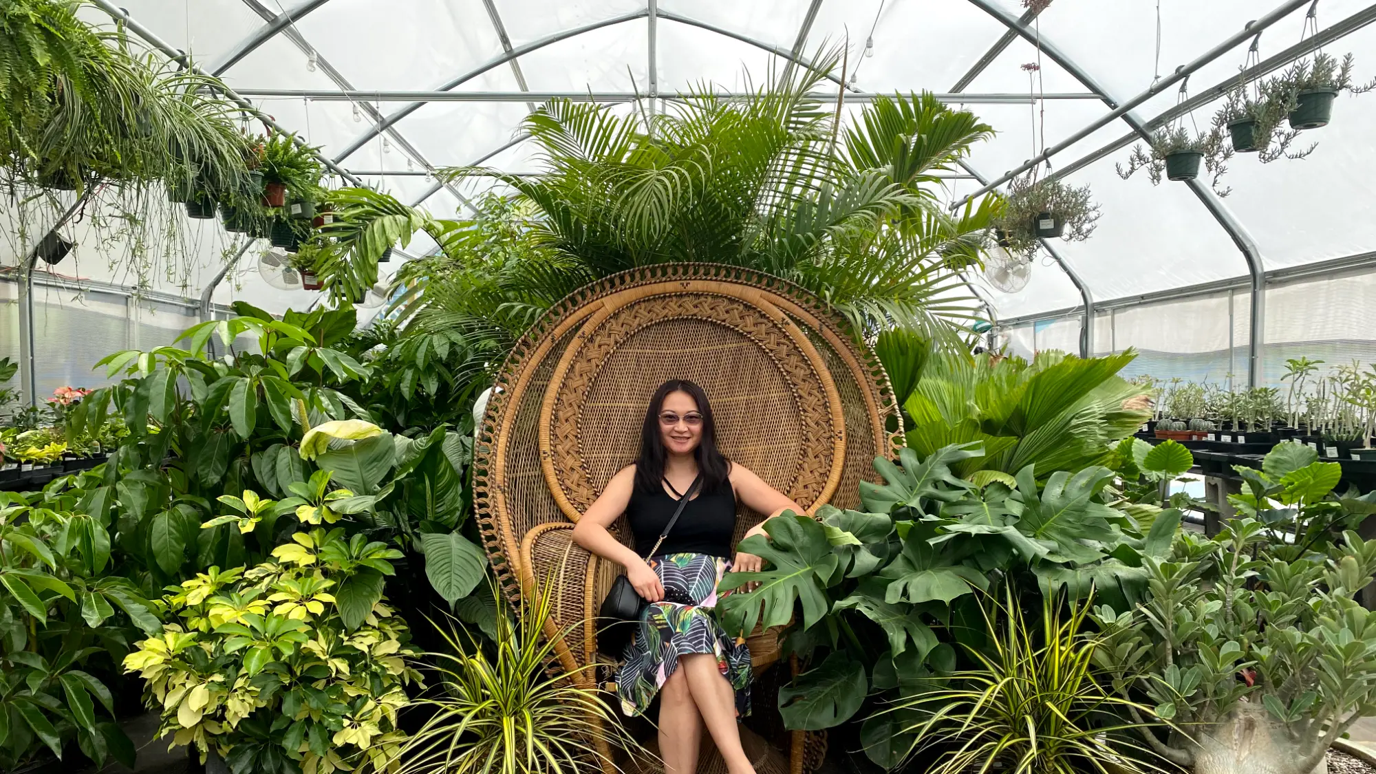 Ellen Hermance seated in a wicker peacock chair surrounded by tropical plants in a greenhouse.